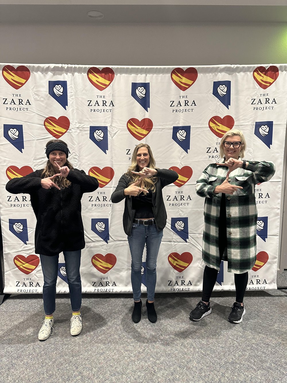 Three community members sharing a playful Z gesture in front of Nevada Vax Watch and The Zara Project signs